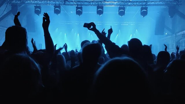 Unrecognizable Fans Dancing and Raising Their Hands at a Concert or Festival Party. Silhouettes of Crowd Audience Enjoying Concert  in Front of Bright Stage Lights.