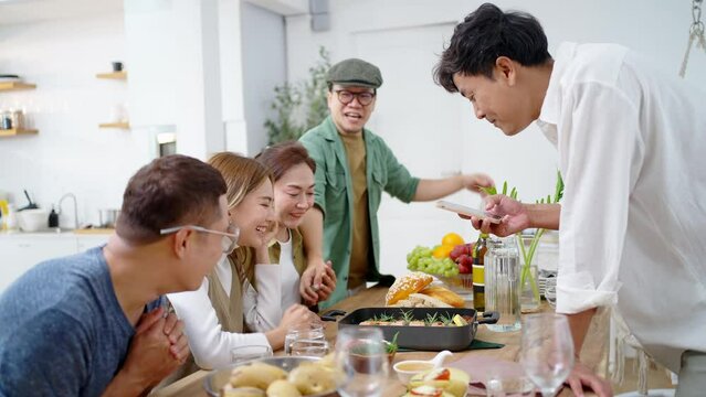 Group of Asian people friends enjoy dinner party and using mobile phone photography food on dining table at home. Happy man and woman reunion meeting celebration together on holiday vacation