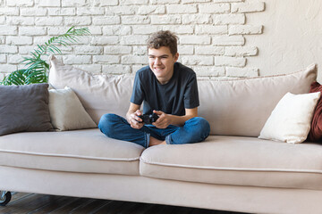 A teenage boy with a wireless joystick in his hands plays a video game sitting at home on the couch. Addiction to games. Free time alone.