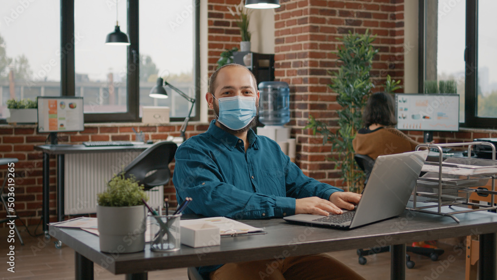 Aufkleber portrait of business man with face mask working with laptop in startup office. employee using comput