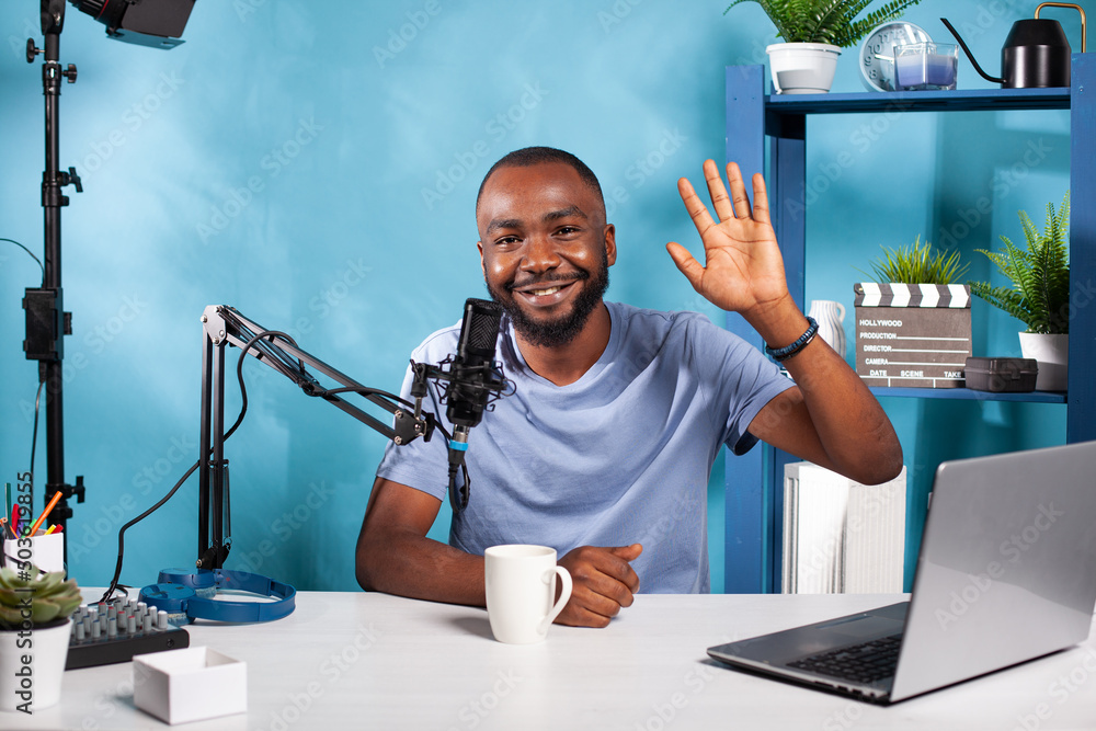 Poster smiling vlogger waving hello sitting at desk in recording studio with professional microphone and vi