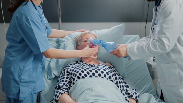 Doctor and nurse using oxygen tube for hyperventilating woman in bed. Medical specialists with tool and heart rate monitor for reanimation and respiratory problem of senior patient.