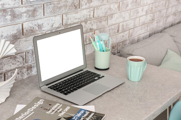 Modern laptop, magazines and cup of tea on table near light brick wall