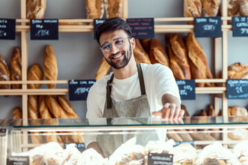 Handsome younger owner selling fresh pastry and loaves un bread section and smiling at pastry shop.