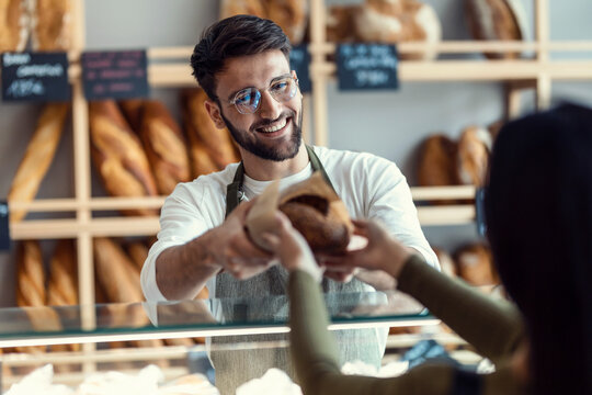 Cheerful seller giving fresh loaf of bread to smiling woman in the pastry shop.
