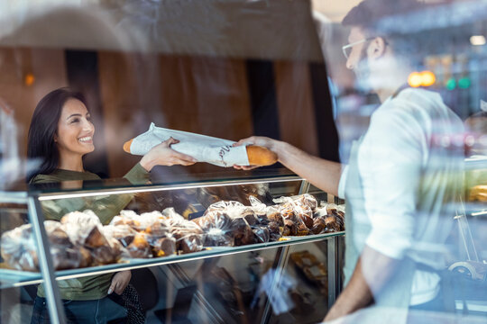 Cheerful seller giving fresh loaf of bread to smiling woman in the pastry shop.