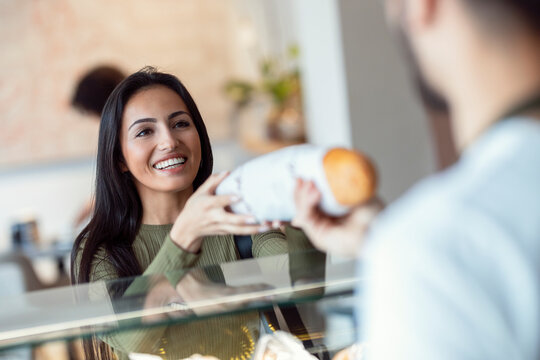 Cheerful seller giving fresh loaf of bread to smiling woman in the pastry shop.