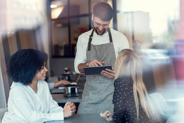 Handsome smiling waitress using application on digital tablet computer to order what the two girls want for breakfast in a pastry shop.