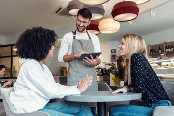 Handsome smiling waitress using application on digital tablet computer to order what the two girls want for breakfast in a pastry shop.