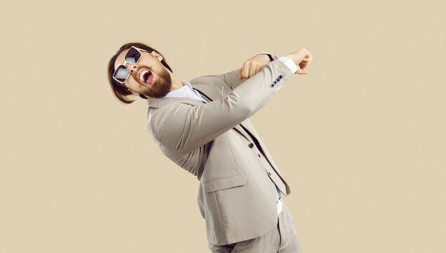 Funny man in suit dancing and having fun. Happy crazy young guy wearing modern beige suit and cool sunglasses dancing and goofing around on beige colour studio background. Fashion and party concept