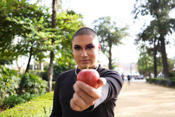 Young non-binary person from South America is showing a red apple to the camera, the person is make up and wearing black clothes and is fat. Concept of health and wellness.