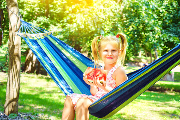Cheerful blonde girl on a hammock eating watermelon in a garden. Fresh and juicy summer snack for a children. Five years old kid enjoying summer.