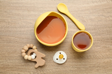 Pacifier, toy and plastic dishware with healthy baby food on wooden table, flat lay