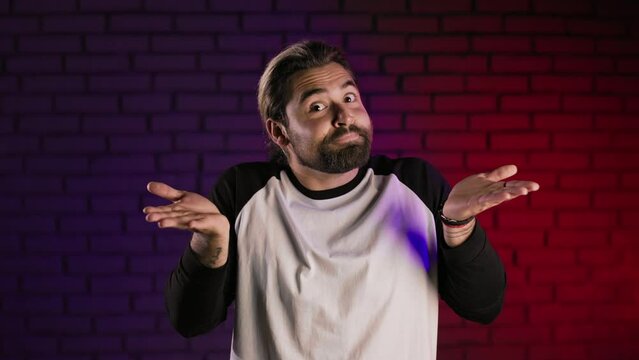 Portrait of confused caucasian man shrugging with shoulders and gesturing with hands while standing in studio with neon light. Male person does not know answer or can not make decision.