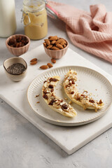 Two banana halves ready with peanut butter topping, raisins, almonds and chia seeds on white plate on marble board