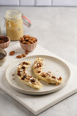 Two banana halves ready with peanut butter topping, raisins, almonds and chia seeds on white plate on marble board