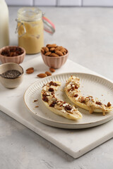 Two banana halves ready with peanut butter topping, raisins, almonds and chia seeds on white plate on marble board