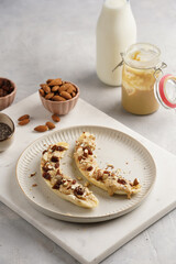 Two banana halves ready with peanut butter topping, raisins, almonds and chia seeds on white plate on marble board