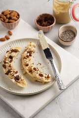 Two banana halves ready with peanut butter topping, raisins, almonds and chia seeds on white plate on marble board