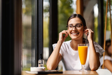 A smiling young woman is sitting in a restaurant and putting wireless earphones on.