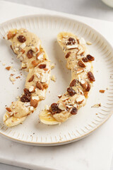 Two banana halves ready with peanut butter topping, raisins, almonds and chia seeds on white plate on marble board