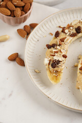 Two banana halves ready with peanut butter topping, raisins, almonds and chia seeds on white plate on marble board