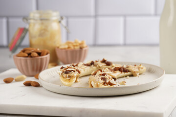Two banana halves ready with peanut butter topping, raisins, almonds and chia seeds on white plate on marble board