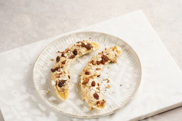 Two banana halves ready with peanut butter topping, raisins, almonds and chia seeds on white plate on marble board, direct creative sunlight shadow