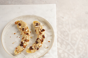 Two banana halves ready with peanut butter topping, raisins, almonds and chia seeds on white plate on marble board, direct creative sunlight shadow. top view