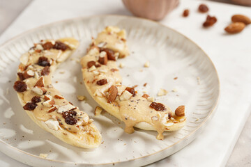 Two banana halves ready with peanut butter topping, raisins, almonds and chia seeds on white plate on marble board, direct creative sunlight shadow