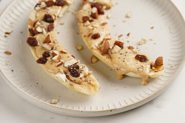 Two banana halves ready with peanut butter topping, raisins, almonds and chia seeds on white plate on marble board, direct creative sunlight shadow