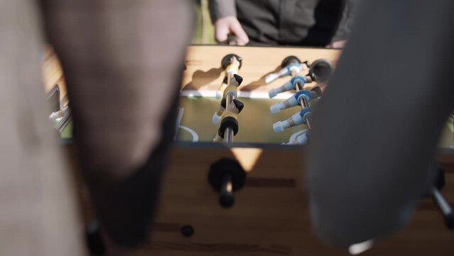 Hands turn handles of table football, plastic figurines of toy football players in blue and yellow colors spin on metal axis and beat off white ball. Men in suits play kicker on sunny lawn, close-up.