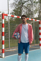 Man holding a soccer ball under his arm. A vertical close-up image of a person in a pink sweatshirt holding a white, red, and blue ball in front of a soccer goal on a blue court with yellow lines.