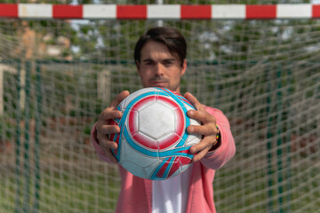Man offering soccer ball to the camera. Close-up image of a person with a pink sweatshirt and white t-shirt showing a white, red and blue ball with both hands on a soccer field.