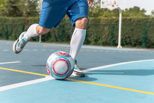 Unrecognizable man shooting the ball on the blue soccer field. Close up image of a young boy wearing blue shorts and white socks kicking a red, white and blue ball.