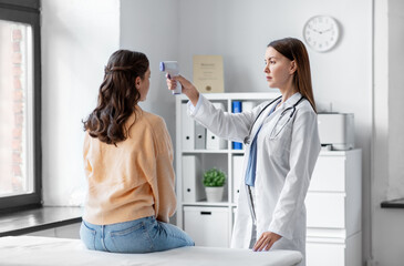 Obraz premium medicine, healthcare and people concept - female doctor with infrared forehead thermometer measuring temperature of woman patient at hospital