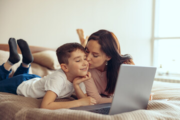 Fototapeta premium Attractive Caucasian mother kissing small teen son lying on bed, looking at laptop screen.