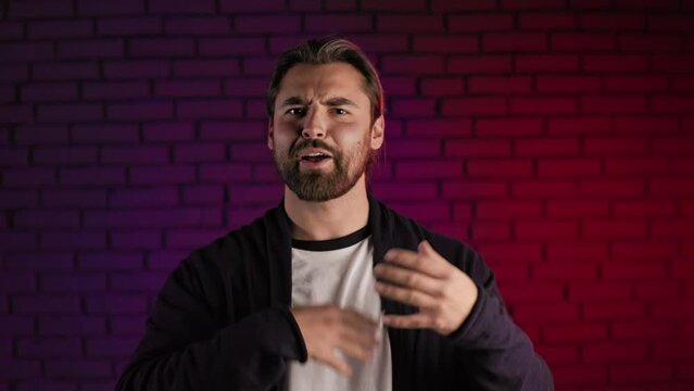 Confident caucasian man in casual talking and gesturing on camera while standing in studio with neon light. Consultation, interview and presentation concept.