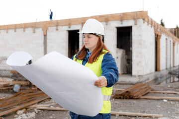 Architect with a blueprints at a construction site. Portrait of successful woman constructor wearing white helmet and safety yellow vest. Woman are planning new project.