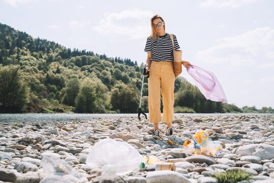 Person help to keep nature clean up and pick up garbage. Young woman clean up outdoor area from rubbish. Volunteer female collecting plastic waste trash on stones of river beach.