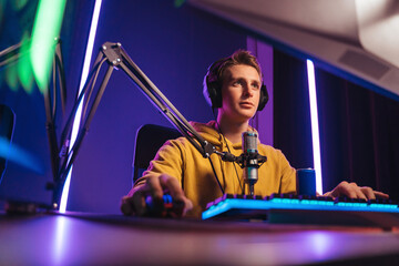 Young professional esport player has live video stream while taking a part in online competition with his team, using microphone and headphones. Competitive gamer playing online video game. Neon light - obrazy, fototapety, plakaty