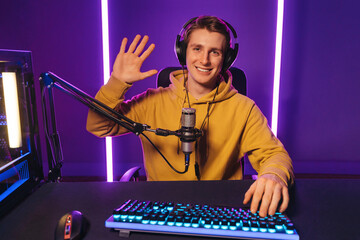 Young handsome vlogger pro gamer waving hand to camera, says hello to his subscribers and followers, smiling on camera. He recording live stream of video game playthrough at home at night. Cyber sport © Arsenii