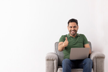 Cheerful young man showing thumb gesture while sitting on sofa with laptop on his lap