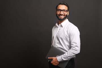 Successful Indian businessman in formal wear and glasses carrying laptop computer standing isolated on black background, portrait of proud hispanic male entrepreneur