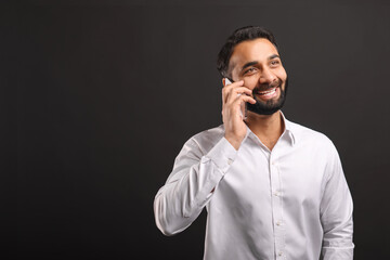 Happy bearded Indian man in formal white shirt talking on smartphone isolated on black background. Male office employee enjoying pleasant phone conversation, chatting on mobile