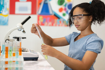 Biracial elementary schoolgirl wearing protective eyewear doing chemical experiment in laboratory - obrazy, fototapety, plakaty