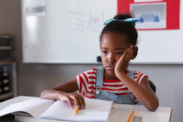 Sad african american girl with hand on cheek and books sitting at desk in classroom