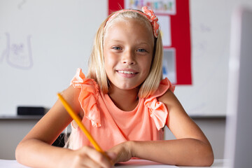 Portrait of smiling caucasian elementary schoolgirl with blond hair studying at desk in classroom