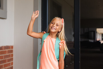 Smiling caucasian elementary schoolgirl waving hand while standing at school entrance