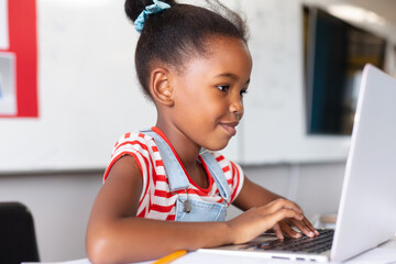 Smiling african american elementary schoolgirl using laptop at desk while studying in class
